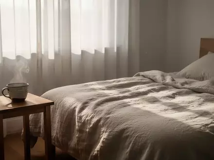 Ein minimalistisch eingerichtetes Schlafzimmer mit einer dampfenden Tasse Wasser auf einem Holztisch im sanften Morgenlicht. - Foto: über boerse-global.de