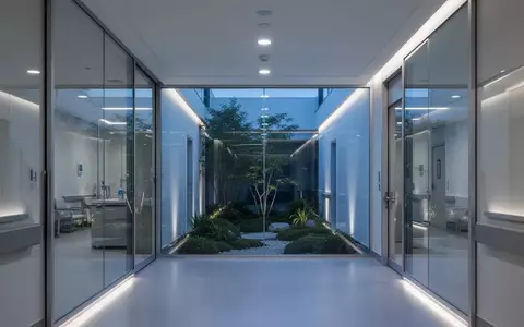Ein moderner, minimalistisch gestalteter Krankenhausflur mit Glasfront und Blick auf einen begrünten Innenhof bei Dämmerung. - Foto: über boerse-global.de