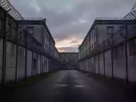 Ein düsterer Gefängnishof mit hohen Betonmauern und Stacheldraht unter einem bewölkten Himmel. - Foto: über boerse-global.de