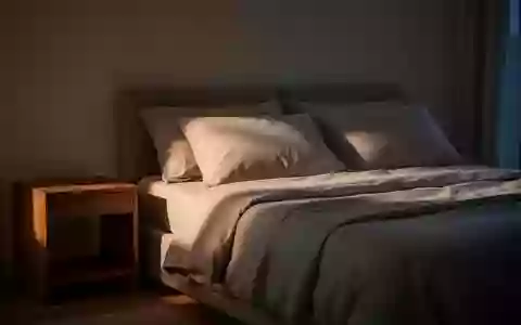 Ein minimalistisch eingerichtetes Schlafzimmer in der Abenddämmerung mit einem ungemachten Bett und einem Holznachttisch. - Foto: über boerse-global.de