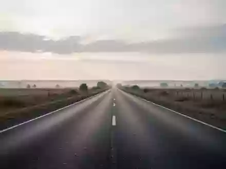 Eine leere, unbefahrene Landstraße erstreckt sich in einer weiten Landschaft unter einem bewölkten Himmel. - Foto: über boerse-global.de