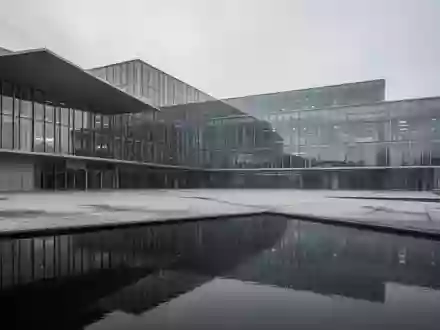 Modernes Verwaltungsgebäude aus Glas und Stahl spiegelt sich in einer Wasserfläche vor einem bewölkten Himmel. - Foto: über boerse-global.de