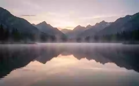 Ein ruhiger Bergsee bei Sonnenaufgang mit spiegelglatter Wasseroberfläche und leichtem Morgennebel. - Foto: über boerse-global.de