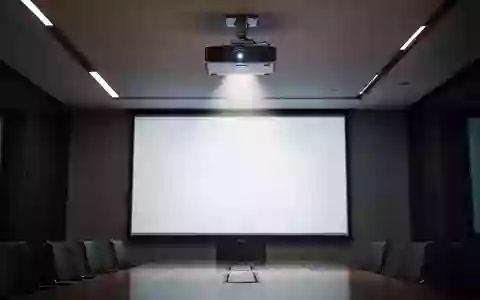 Ein moderner, abgedunkelter Konferenzraum mit einem Deckenprojektor, der auf eine leere, weiße Leinwand strahlt. - Foto: über boerse-global.de