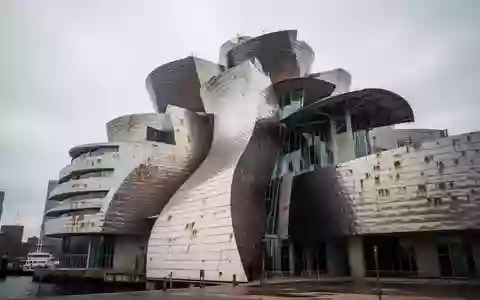 Die geschwungene, metallisch glänzende Fassade der Düsseldorfer Gehry-Bauten vor einem bewölkten Himmel. - Foto: über boerse-global.de