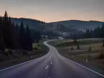 Eine einsame, kurvige Landstraße, die sich durch eine hügelige Waldlandschaft bei Dämmerung schlängelt. - Foto: über boerse-global.de