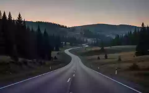 Eine einsame, kurvige Landstraße, die sich durch eine hügelige Waldlandschaft bei Dämmerung schlängelt. - Foto: über boerse-global.de