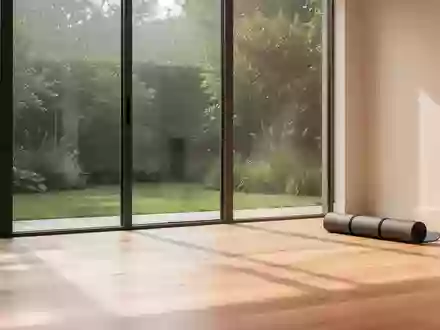 Ein minimalistischer, lichtdurchfluteter Übungsraum mit Holzboden und einer zusammengerollten Yogamatte vor einem Gartenblick. - Foto: über boerse-global.de