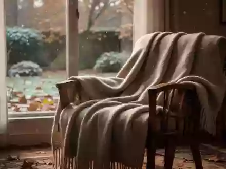 Eine gemütliche, warm beleuchtete Wohnzimmerecke mit einer weichen Wolldecke auf einem Sessel vor einem Fenster an einem Herbsttag. - Foto: über boerse-global.de