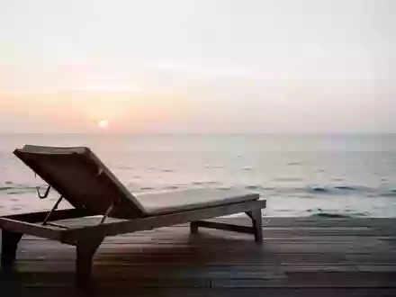 Ein leerer Liegestuhl auf einer Holzterrasse mit Blick auf das ruhige Meer bei Sonnenaufgang. - Foto: über boerse-global.de