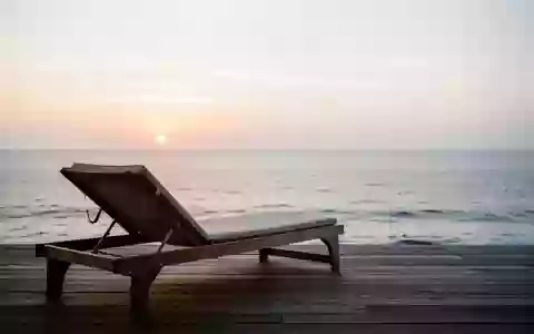 Ein leerer Liegestuhl auf einer Holzterrasse mit Blick auf das ruhige Meer bei Sonnenaufgang. - Foto: über boerse-global.de
