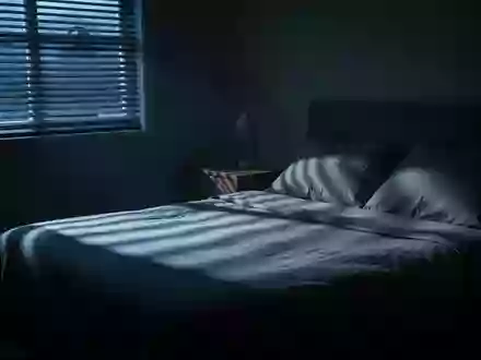 Ein ruhiges, abgedunkeltes Schlafzimmer im Morgengrauen mit sanftem Lichteinfall und langen Schatten auf dem unberührten Bett. - Foto: über boerse-global.de