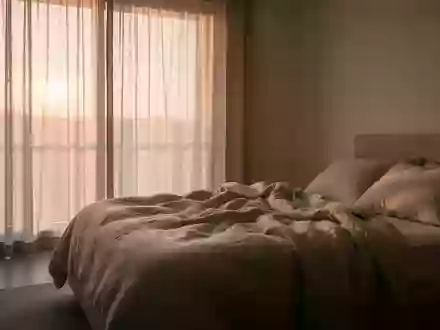 Ein minimalistisch eingerichtetes Schlafzimmer in ruhigem Morgenlicht mit ungemachtem Bett und neutralen Textilien. - Foto: über boerse-global.de