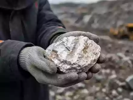 Ein Bergbauingenieur hält ein rohes Lithium-Mineral in den Händen vor einem unscharfen Industriegelände. - Foto: über boerse-global.de