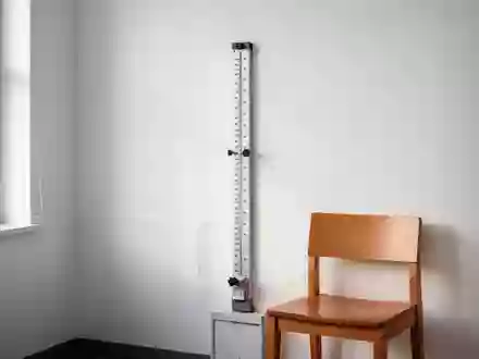 Ein minimalistisch eingerichtetes medizinisches Untersuchungszimmer mit einem Messstab an einer weißen Wand und einem leeren Stuhl. - Foto: über boerse-global.de