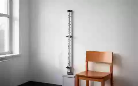 Ein minimalistisch eingerichtetes medizinisches Untersuchungszimmer mit einem Messstab an einer weißen Wand und einem leeren Stuhl. - Foto: über boerse-global.de