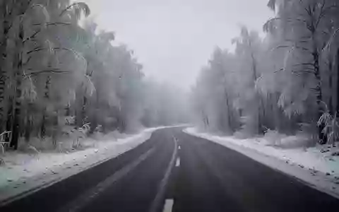 Eine verschneite, menschenleere Landstraße in einem frostigen Wald bei trübem Winterwetter. - Foto: über boerse-global.de