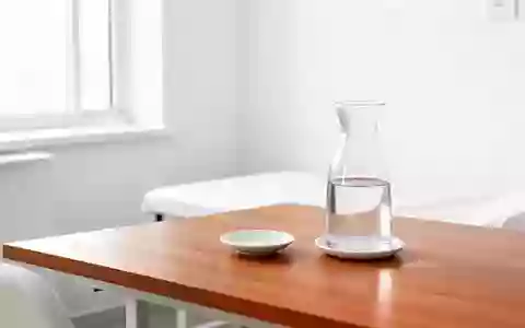 Ein minimalistisch eingerichteter, heller Behandlungsraum mit einer Wasserkaraffe und einer Untertasse auf einem Holztisch. - Foto: über boerse-global.de