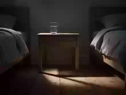 Ein schlichtes Nachttischchen mit einem Glas Wasser in einem abgedunkelten Schlafzimmer bei nächtlichem Mondlicht. - Foto: über boerse-global.de
