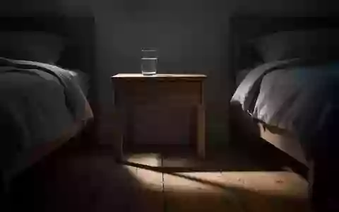Ein schlichtes Nachttischchen mit einem Glas Wasser in einem abgedunkelten Schlafzimmer bei nächtlichem Mondlicht. - Foto: über boerse-global.de