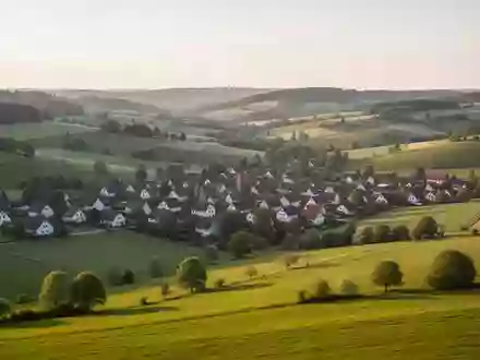 Weitläufige Landschaft mit Wiesen und Feldern am Rande einer Ortschaft in einer hügeligen Region bei Sonnenuntergang. - Foto: über boerse-global.de