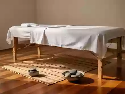 Eine schlichte Massageliege mit weißen Laken in einem hellen, ruhigen Raum mit Bambusmatte und glatten Steinen. - Foto: über boerse-global.de