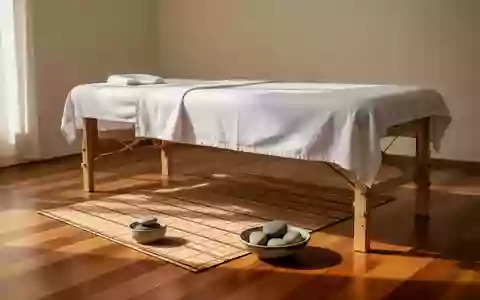 Eine schlichte Massageliege mit weißen Laken in einem hellen, ruhigen Raum mit Bambusmatte und glatten Steinen. - Foto: über boerse-global.de