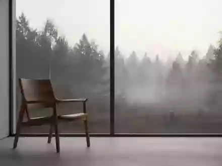 Ein minimalistischer, ruhiger Raum mit einem leeren Holzstuhl vor einem großen Fenster, das den Blick auf einen nebligen Wald freigibt. - Foto: über boerse-global.de