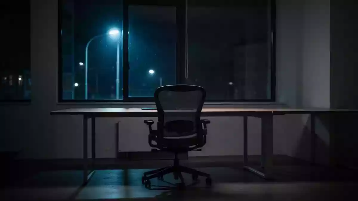 Ein dunkler, moderner Arbeitsplatz bei Nacht mit einem leeren Schreibtisch, der durch das schwache Licht eines Straßenfensters beleuchtet wird. Illustration mit AI erstellt.