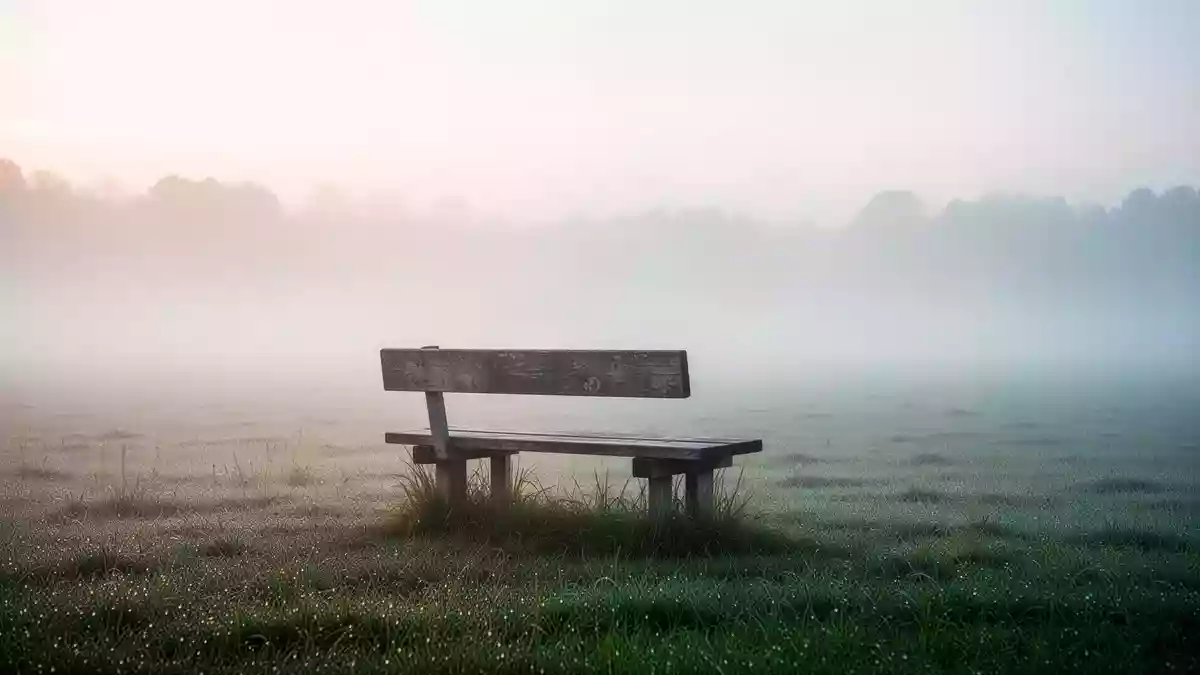 Eine einsame, verwitterte Holzbank auf einer nebligen Wiese bei Sonnenaufgang, die Ruhe und Reduktion ausstrahlt. Illustration mit AI erstellt.