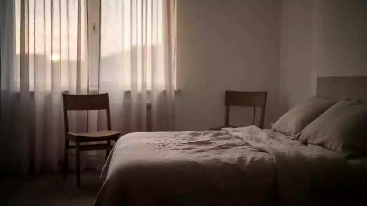 Ein ruhiges, minimalistisches Schlafzimmer im sanften Licht der Morgendämmerung mit einem leeren Stuhl am Fenster. Illustration mit AI erstellt.