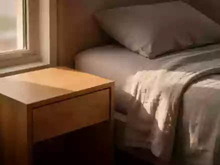 Ein minimalistischer Nachttisch in einem hellen, ruhigen Schlafzimmer bei Sonnenaufgang mit weichem Licht auf dem Bett. - Foto: über boerse-global.de