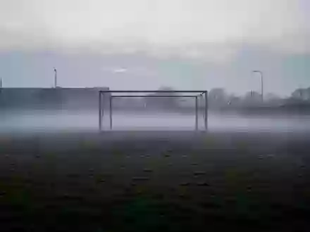 Ein leeres, nebliges Fußballfeld in der Dämmerung mit einem metallenen Torpfosten im Hintergrund. - Foto: über boerse-global.de