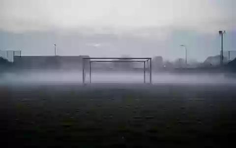 Ein leeres, nebliges Fußballfeld in der Dämmerung mit einem metallenen Torpfosten im Hintergrund. - Foto: über boerse-global.de