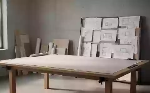 Ein minimalistischer, heller Design-Arbeitsbereich mit einem leeren Zeichentisch und Architekturplänen an einer Betonwand. - Foto: über boerse-global.de