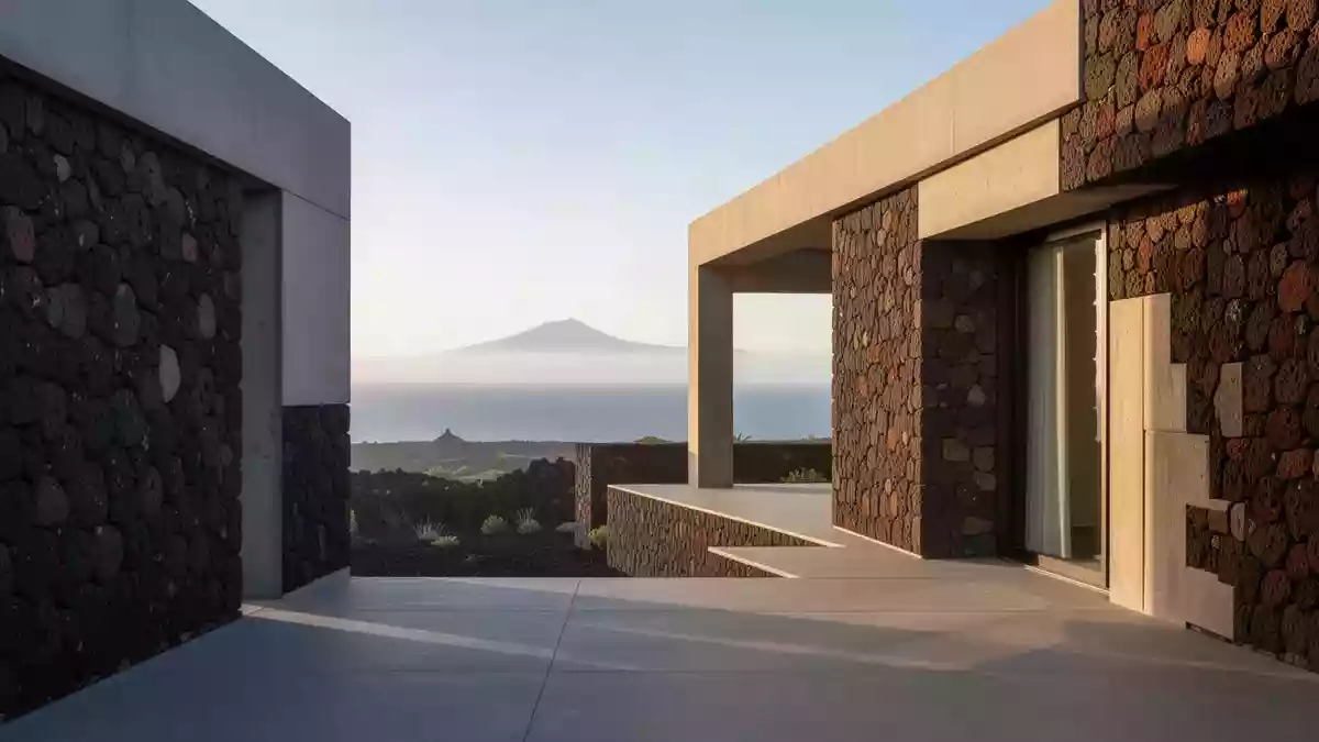 Eine moderne Architekturfassade aus Vulkangestein vor einer weitläufigen Küstenlandschaft auf Teneriffa bei Morgenlicht. Illustration mit AI erstellt.