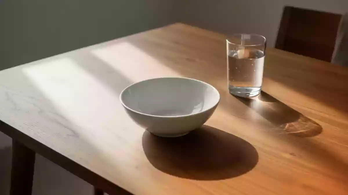 Ein leerer weißer Keramikteller und ein Glas Wasser auf einem hölzernen Esstisch im natürlichen Morgenlicht. Illustration mit AI erstellt.