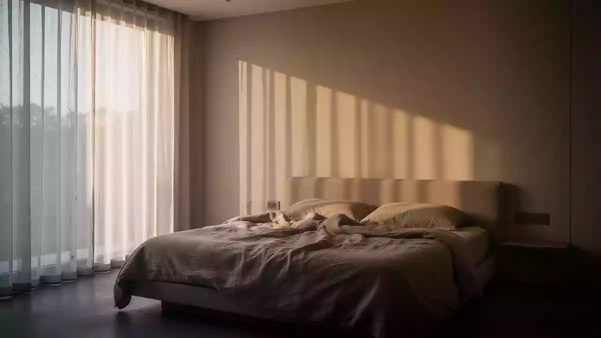 Ein minimalistisch eingerichtetes Schlafzimmer am frühen Morgen mit sanftem, durch Vorhänge einfallendem Tageslicht. Illustration mit AI erstellt.