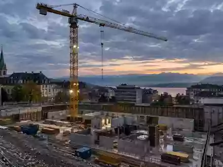 Baustelle in einer Schweizer Stadt mit einem Turmdrehkran vor einem bewölkten Himmel bei Dämmerung. - Foto: über boerse-global.de
