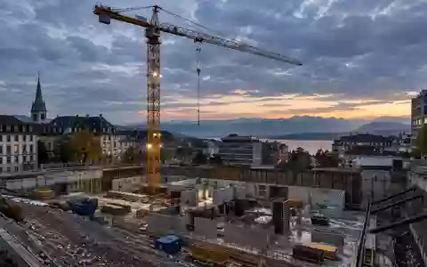 Baustelle in einer Schweizer Stadt mit einem Turmdrehkran vor einem bewölkten Himmel bei Dämmerung. - Foto: über boerse-global.de