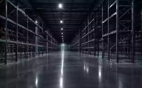 Weitläufiges, leeres Logistiklager mit hohen Metallregalen und reflektierendem Betonboden in einer kühlen, sterilen Atmosphäre. - Foto: über boerse-global.de