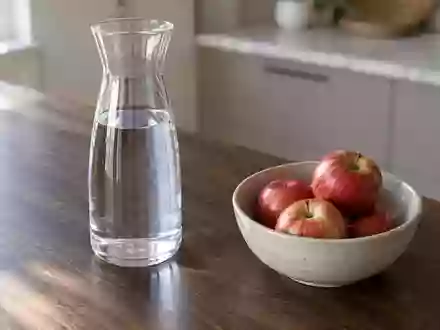 Ein Glas Wasser und eine Schale mit frischen Äpfeln auf einem Holztisch in einer hellen Küche. - Foto: über boerse-global.de