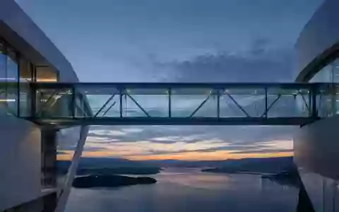 Eine moderne Glas-Stahl-Brücke bei Dämmerung, die zwei abstrakte architektonische Strukturen verbindet. - Foto: über boerse-global.de