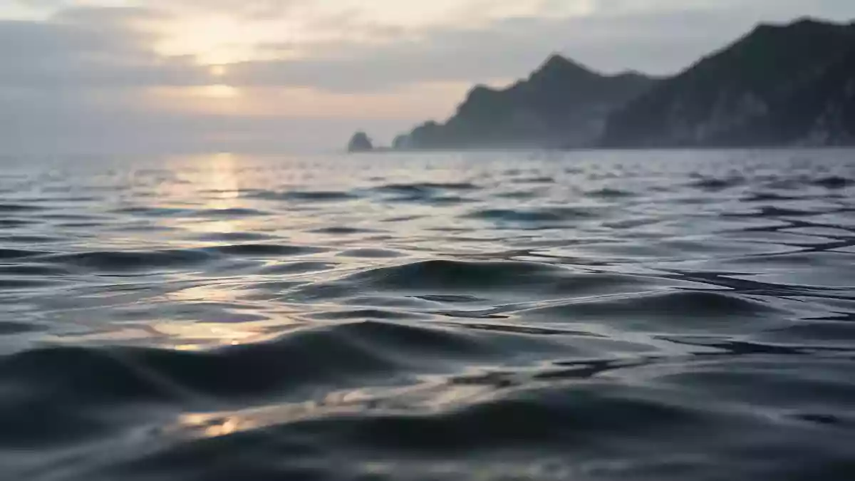 Nahaufnahme einer ruhigen Wasseroberfläche mit sanften Wellen und weichem Licht, im Hintergrund eine verschwommene Küstenlinie. Illustration mit AI erstellt.