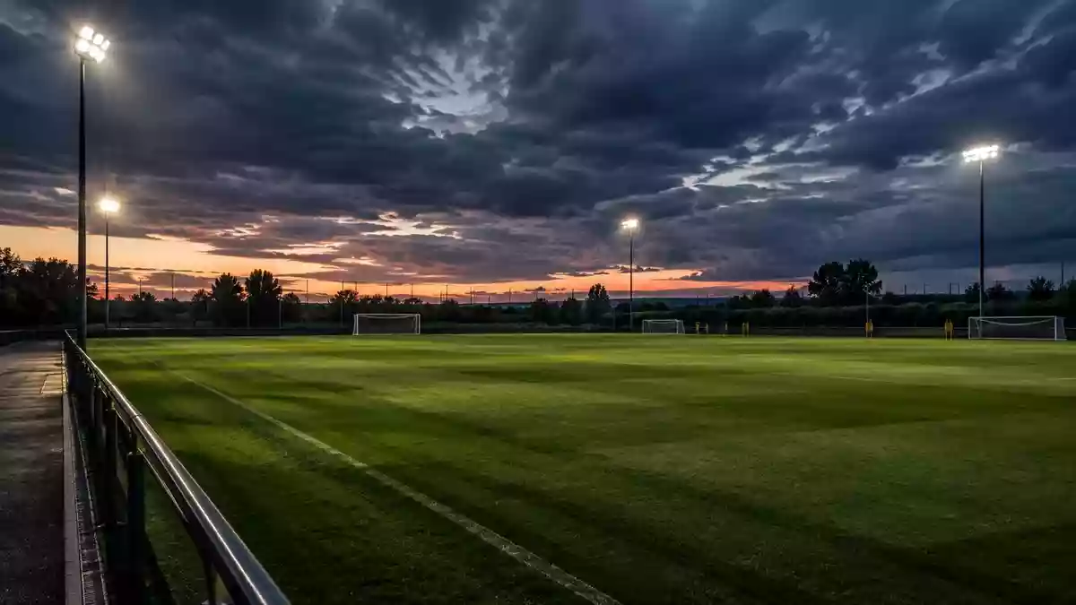 Ein leerer Fußballtrainingsplatz bei Dämmerung mit langen Schatten auf dem Rasen und dramatischem Himmel. Illustration mit AI erstellt.