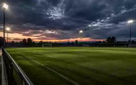 Ein leerer Fußballtrainingsplatz bei Dämmerung mit langen Schatten auf dem Rasen und dramatischem Himmel. - Foto: über boerse-global.de