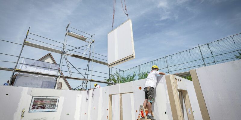 Ein Bauarbeiter steht auf einer Leiter und arbeitet im Rohbau eines Fertighauses, während gerade ein weiteres Wandelement von einem Kran heruntergelassen wird. - Foto: Daniel Maurer/dpa