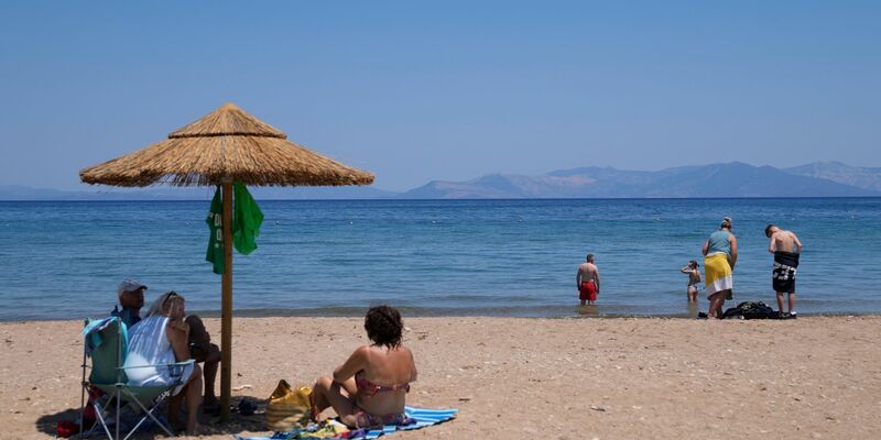 Da helfen nur noch Sonnenschrirme und ein Handventilator: Eine Frau am Strand von Loutraki. - Foto: Thanassis Stavrakis/AP/dpa