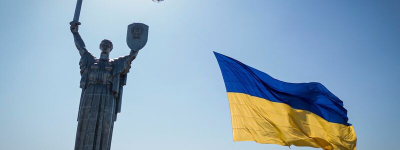 Eine Drohne trägt eine große Nationalflagge vor das ukrainische Mutterland-Denkmal in Kiew - Foto: Evgeniy Maloletka/AP/dpa