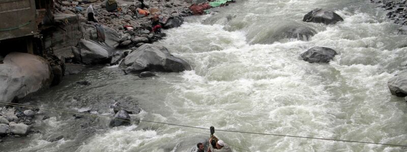 Menschen überqueren einen Fluss mithilfe einer Seilbahn. - Foto: Naveed Ali/AP/dpa